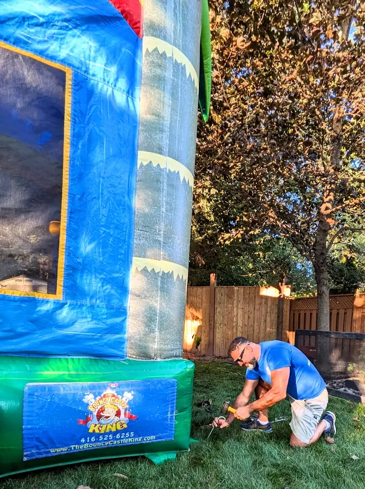 The Bouncy Castle King team member hammering a ground stake beside a bouncy castle on grass during a Brampton-area setup